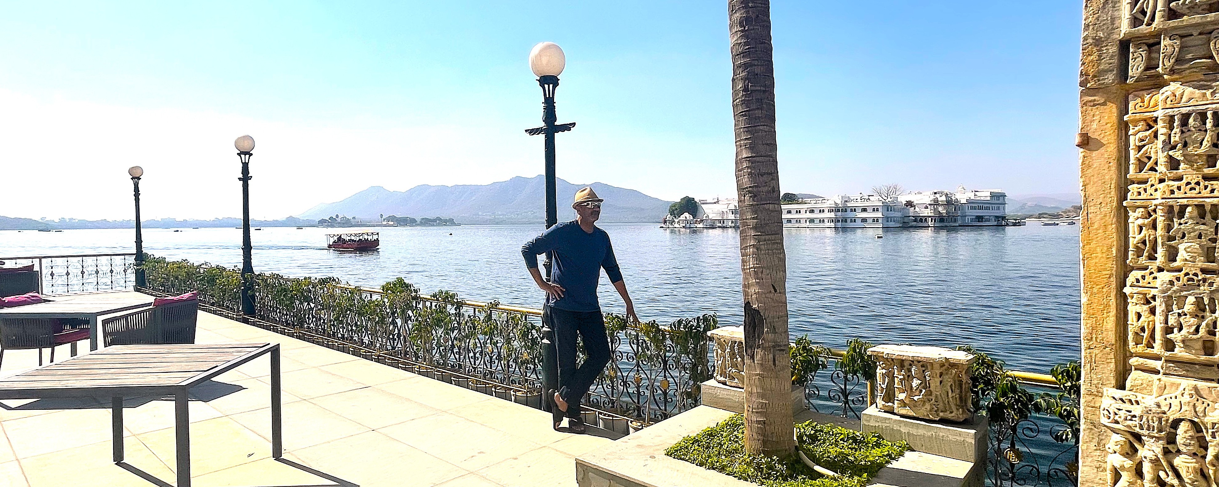 A traveler on a lakeside terrace overlooking a white palace and distant hills