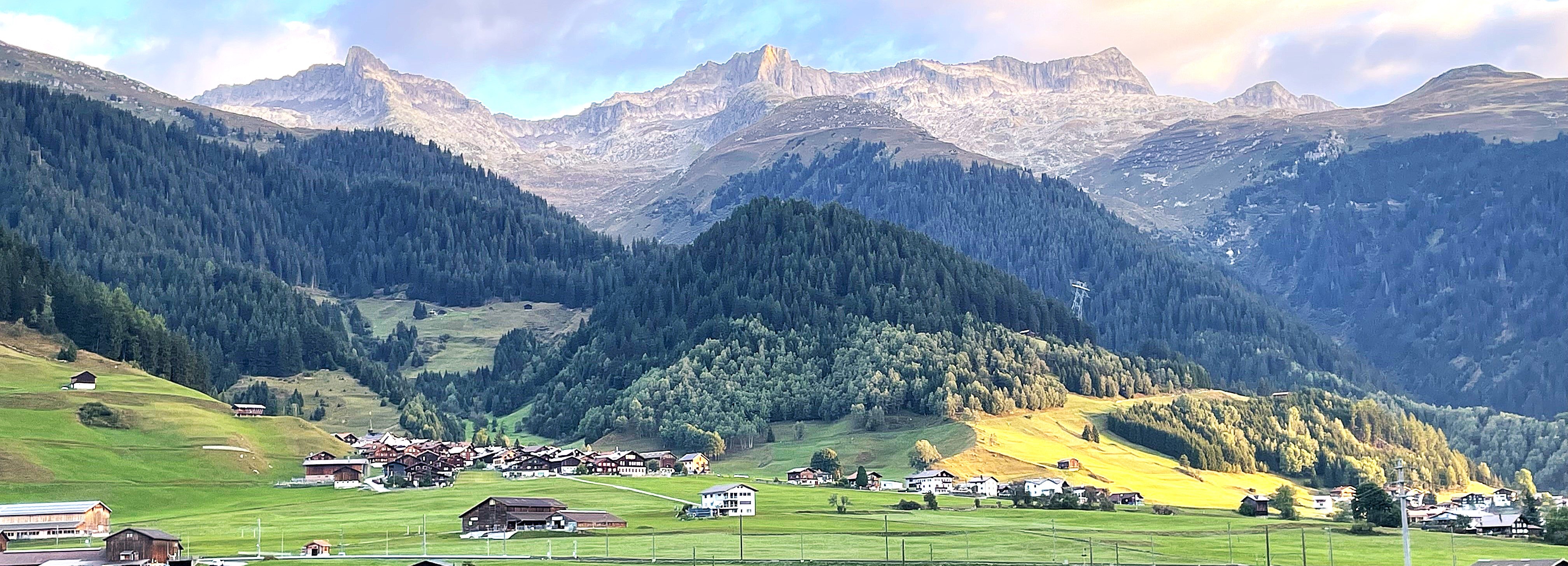 An Alpine village surrounded by green meadows, forested hills and mountain peaks