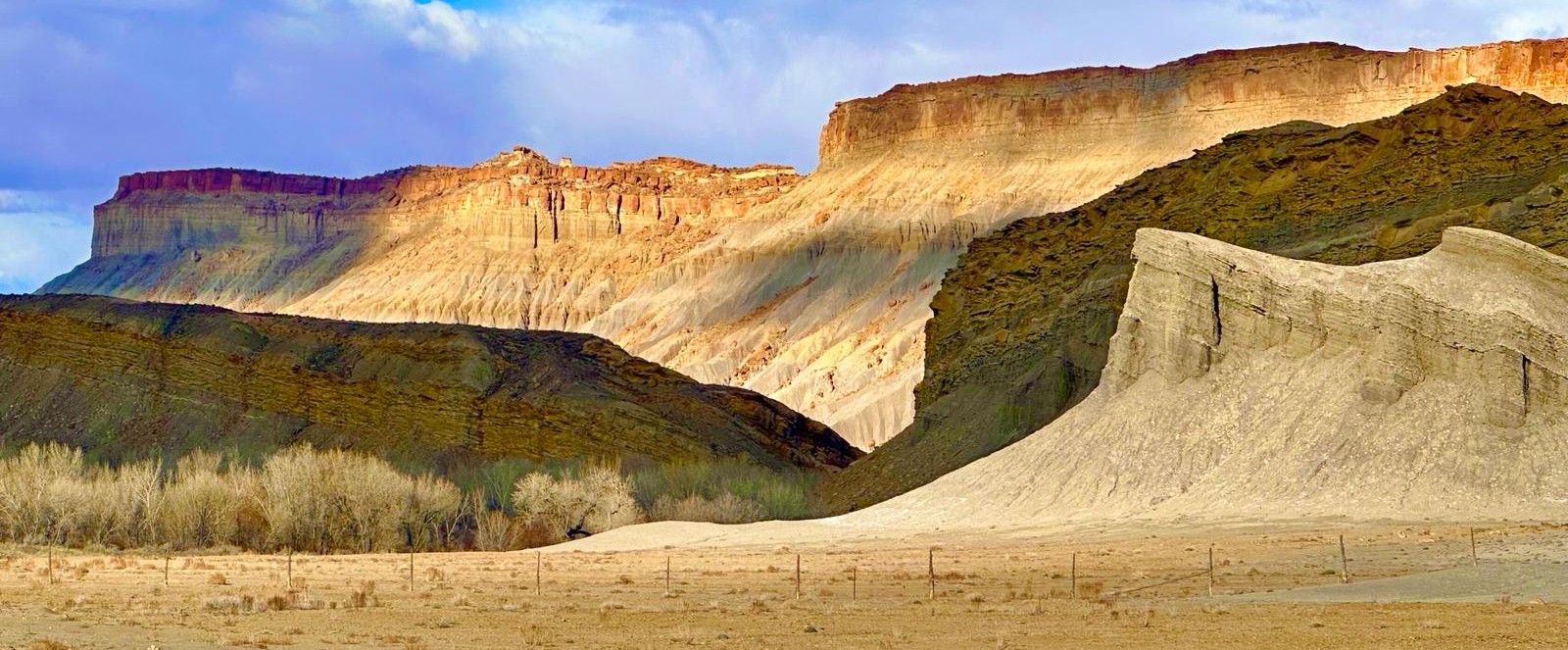 Sunlit desert cliffs and layered rock formations beneath a blue, clouded sky